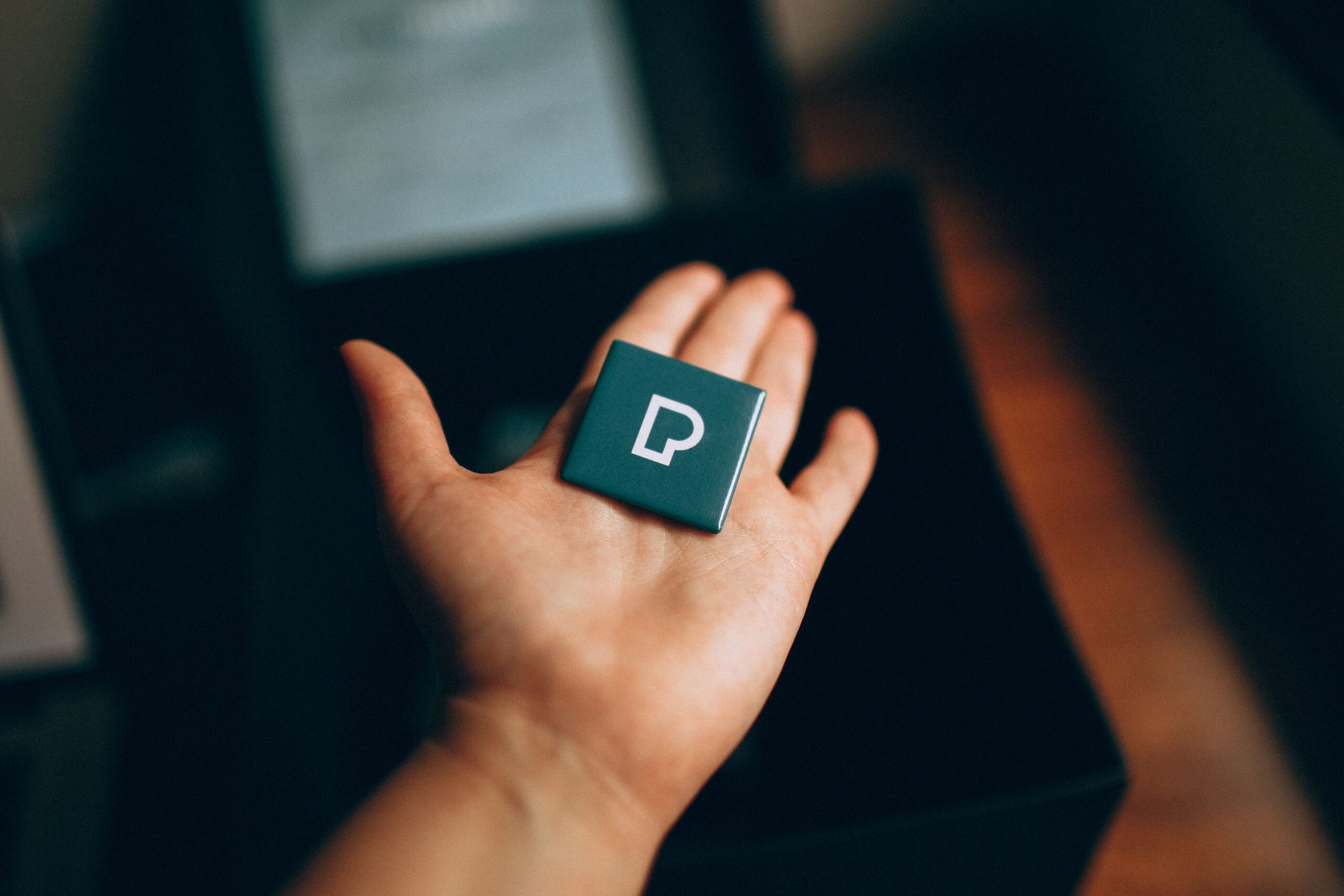 An open hand presenting a square pin with a letter 'P' design in a dark setting.